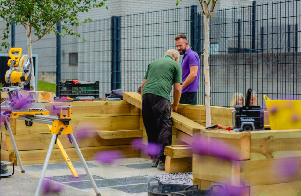 Two gardeners construct a raised wooden planter in an outdoor space, surrounded by tools, materials, and flowering plants.