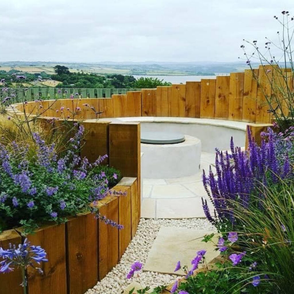 A beautifully landscaped garden with curved wooden seating, a central fire pit, and lush purple flowers, overlooking a scenic countryside view.
