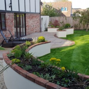 A neatly landscaped garden features curving white-raised beds with brick edging, a lush lawn, a paved patio with reclining chairs, and a tranquil, sunny atmosphere.