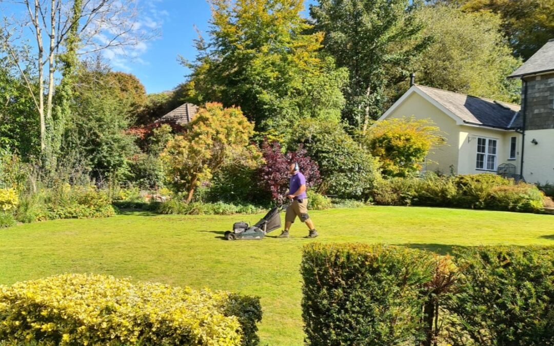 Gardener cutting grass in a large lawned garden, surrounded by trees and bushes, with part of a house visible in the background.