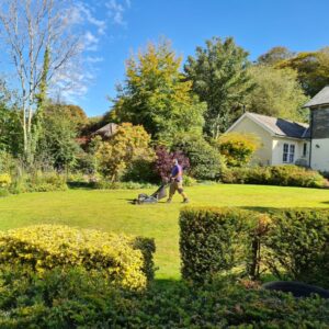 Gardener cutting grass in a large lawned garden, surrounded by trees and bushes, with part of a house visible in the background.