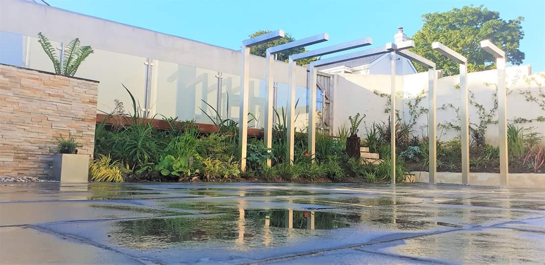 A modern landscaped garden featuring a wet paved patio, raised planting beds with lush greenery, and sleek metal pergolas against a white wall with climbing plants.